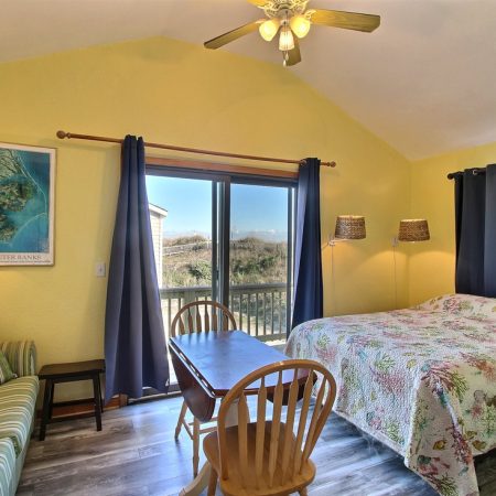 Sunny bedroom with a double bed, small table, two chairs, sofa, map on wall, and sliding glass doors to a balcony.