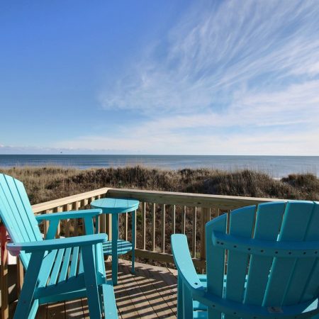 Two blue Adirondack chairs on a wooden deck overlook a sandy beach and calm ocean under a partly cloudy sky.
