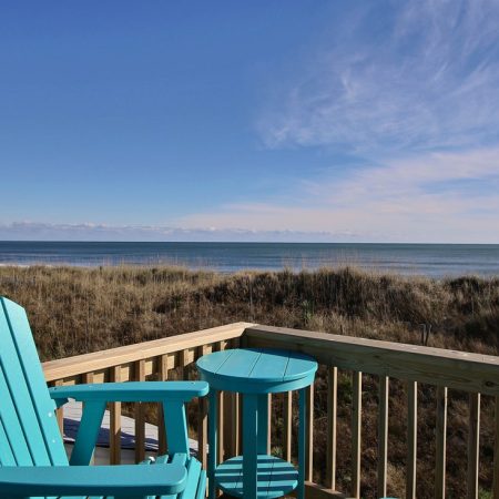 Turquoise chair and table on a wooden deck overlook sand dunes and the ocean under a clear blue sky.