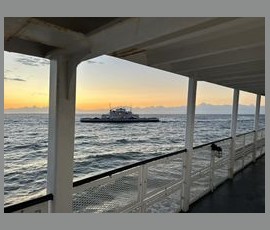 View of a ferry on the water at sunset, seen from the deck of another boat with metal railings and a covered roof.