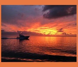 A boat moves across calm water at sunset under a sky filled with orange and pink clouds.