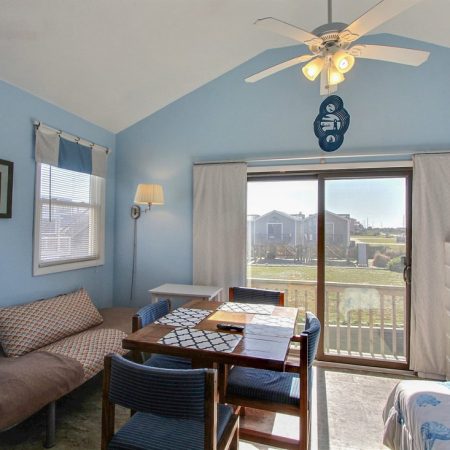 Small blue-themed room with a table, chairs, futon, bed, fridge, and sliding glass door leading to a balcony.