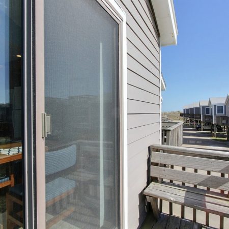 Small wooden balcony with glass sliding door, next to gray house siding; row of raised houses visible in background.