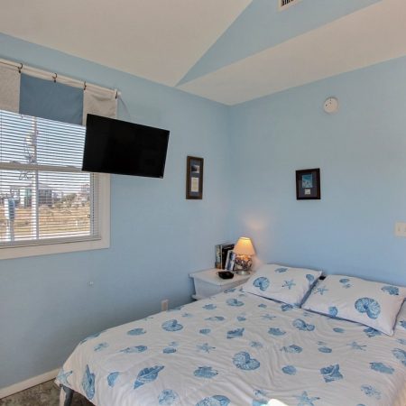 Small bedroom with a bed, blue seashell bedding, wall-mounted TV, nightstand, lamp, and window with blinds.