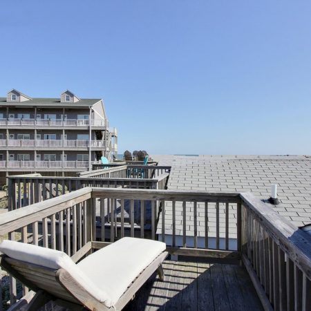 Wooden deck with a lounge chair overlooks rooftops, apartment building, and a clear, blue sky in the background.