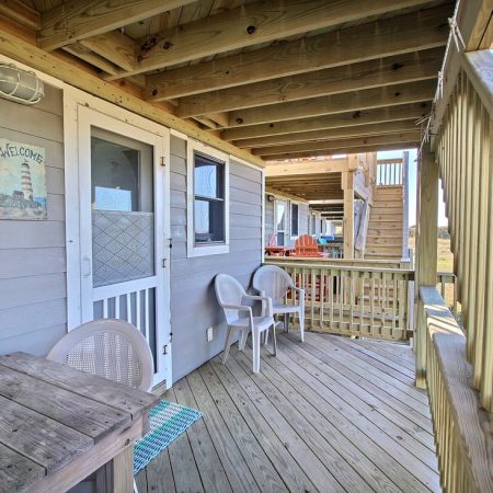 Wooden porch with a small table, two chairs, stairs, and a white door. Wall sign says “Welcome” with a lighthouse.