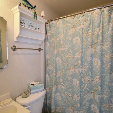 Bathroom with a blue ocean-themed shower curtain, white shelves above toilet, and a small sink at left.