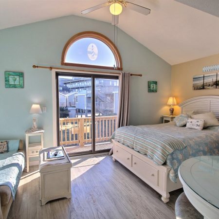 Bright bedroom with a queen bed, sliding glass doors to a balcony, sofa, TV, and beach-themed decor.