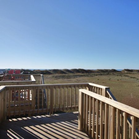 Wooden deck with railings overlooks grassy dunes and distant beach houses under a clear blue sky.