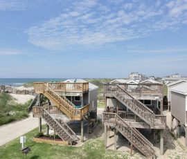 Two elevated beach houses with wooden staircases, situated near sand dunes and the ocean under a partly cloudy sky.