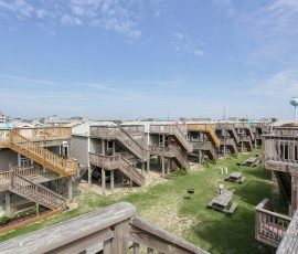 Row of raised beach houses with wooden stairs and picnic tables on grassy area under a partly cloudy sky.