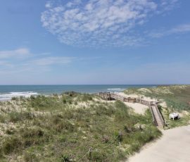 Wooden boardwalk over sandy dunes leads to a beach with grassy patches under a blue sky with scattered clouds.