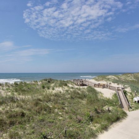 Wooden boardwalk crosses sandy dunes, leading to the beach and ocean under a partly cloudy blue sky.