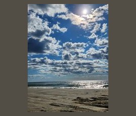 Sunny beach scene with a bright sky, scattered clouds, and sunlight reflecting on the ocean waves.