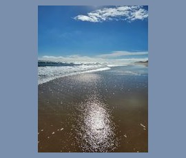 Waves gently washing onto a sandy beach under a bright blue sky with scattered clouds and sunlight reflecting on the water.