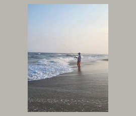 Person fishing while standing at the edge of the ocean on a sandy beach under a clear sky.