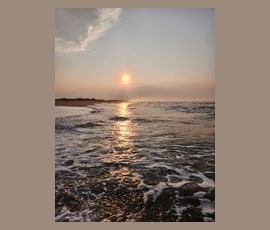 Sunset over the ocean with waves approaching the shore, reflecting sunlight on the water’s surface.