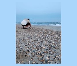 A person squats on a shell-covered beach near the ocean, looking down at the sand under a cloudy sky.