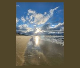 Sunlight reflects off wet sand on a beach under a partly cloudy sky, with gentle waves along the shoreline.