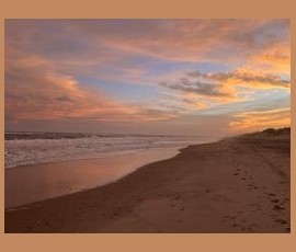Sunset over a sandy beach with gentle waves and colorful clouds in the sky. Footprints are visible along the shoreline.