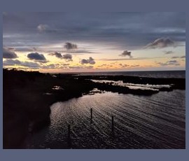 Coastline at dusk with dark clouds, calm waves, and wooden posts in the water near the shore.