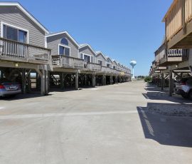 Row of elevated beach houses with wooden balconies and parked cars underneath, on a sunny day.