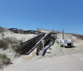 Wooden ramps and stairs lead over sand dunes to a beach under a clear blue sky, with a rinse station nearby.