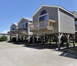 Three gray beach houses on stilts with wooden balconies, set along a paved driveway under a clear blue sky.