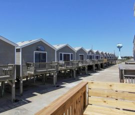 Row of gray, elevated beach cottages with wooden decks under a clear blue sky and a water tower in the background.