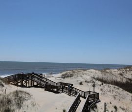 Wooden boardwalk over sand dunes leads to a calm beach with blue sky and gentle waves in the background.
