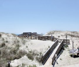 Wooden walkways cross sandy dunes under a clear sky; a person sits at a bench near the entrance.