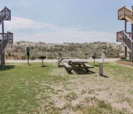 Two picnic tables and grills on grassy area between two wooden staircases, with sand dunes in the background.