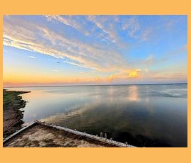 Calm water stretches to the horizon under a colorful sky at sunrise, with a concrete edge in the foreground.