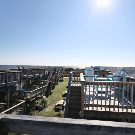 Wooden decks and staircases overlook grassy area, facing the ocean under a clear blue sky with bright sunlight.