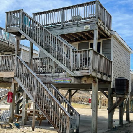 Elevated beach house with exterior wooden stairs, railings, and a covered area underneath, on a sunny day.