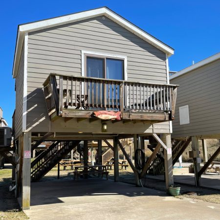Elevated gray beach house with a small balcony, wooden stairs, and open parking space underneath on a clear day.
