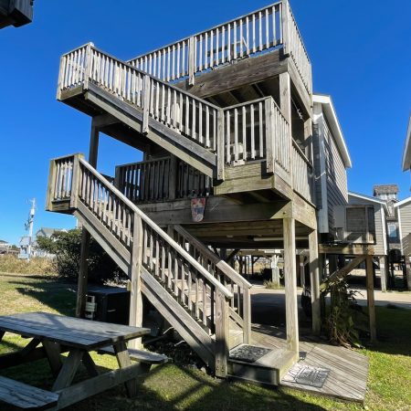 Elevated wooden beach house with two staircases, wraparound decks, and a picnic table on the grass below.