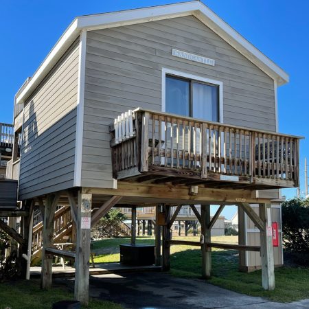 Beige stilt beach house with a small balcony, wooden stairs, and clear blue sky in the background.