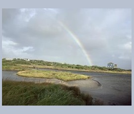 A faint rainbow arcs over grassy dunes and a blue house near a calm body of water under a cloudy sky.