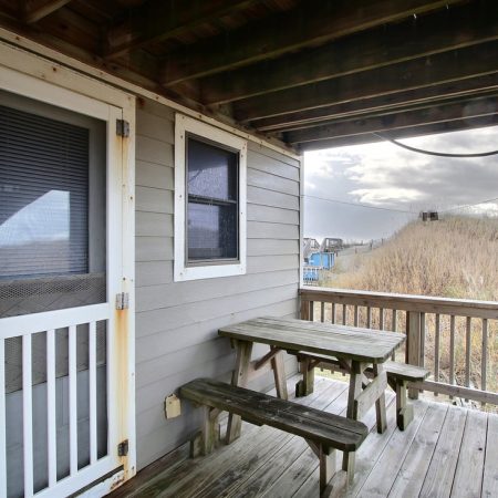 Small porch with wooden picnic table and benches, screened door, window, and view of grassy area under cloudy sky.