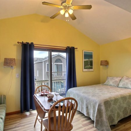 Small bedroom with a bed, striped couch, dining table, ceiling fan, and window with blue curtains.