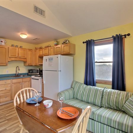 Small kitchen and dining area with a striped sofa, wooden table, and light wood cabinets under a bright window.