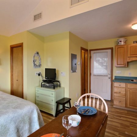 Small studio apartment with a bed, kitchenette, dining table, and TV on a dresser, featuring light yellow walls.