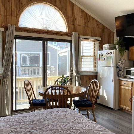 Small kitchen and dining area with wooden cabinets, round table, large windows, and a bed in the foreground.