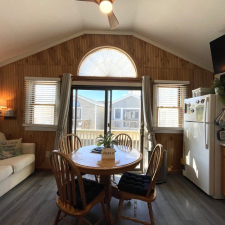 Dining area with round table, four chairs, large window, refrigerator, and wooden wall accents in a cozy kitchen space.