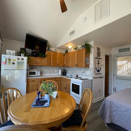 Small kitchen with wood cabinets, dining table, and bed in bright, cozy room; plants and nautical decor visible.