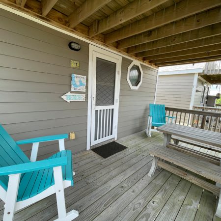 Wooden porch with turquoise chairs, a bench table, gray siding, and a white door under a covered deck.