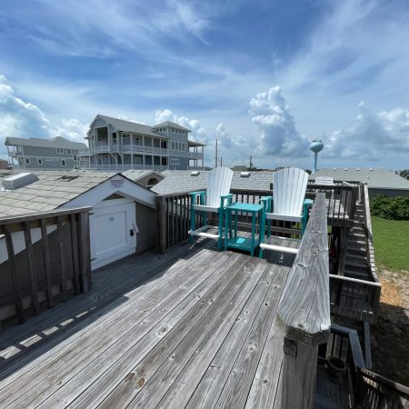 Two white Adirondack chairs and a small table sit on a wooden rooftop deck under a partly cloudy sky.