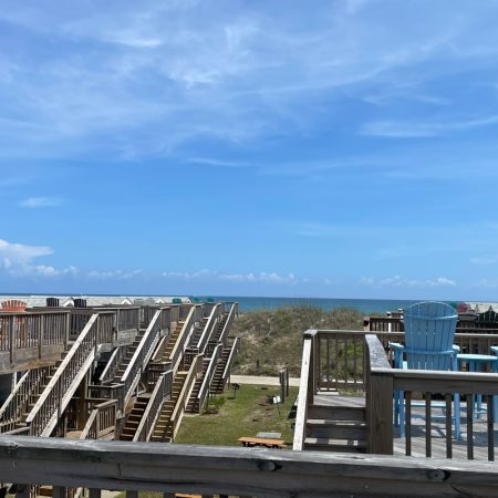 Wooden decks and staircases overlook grassy dunes and the ocean under a blue sky with scattered clouds.