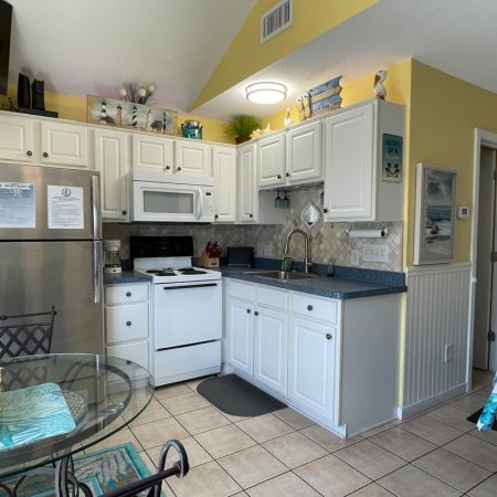 Small kitchen with white cabinets, stainless refrigerator, stove, microwave, sink, glass table, and yellow walls.